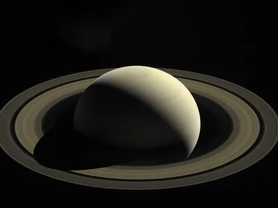 The smooth surface of a round planet is partially illuminated by the sun; the planet's other half is in shadow. The planet is surrounded by thousands of rings.