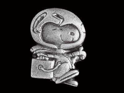 A small silver pin shows a smiling Snoopy, a cartoon dog, wearing a spacesuit with a large clear glass helmet, performing his iconic happy beagle dance.