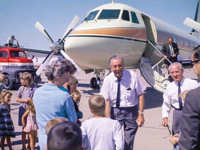 Walt Disney, an senior man in a white button up short-sleeved shirt, with a tie and slacks, greets a small crowd of people in front of his airplane, which is large enough it extends out of frame and is white with orange and blue stripes.