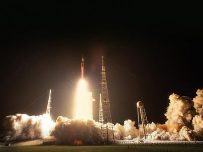 The pitch black night is illuminated by a bright white-hot rocket engine plume, which illuminates clouds of smoke surrounding the launch pad.