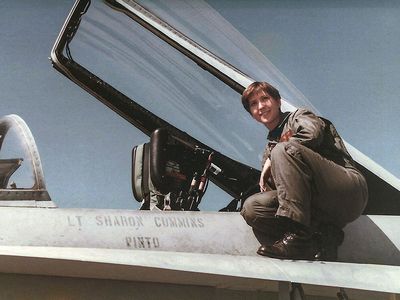 A young woman wearing a U.S. Navy olive-green flightsuit and black combat boots perches next to the cockpit of her F/A-18C miltary jet. "Lt Sharon Cummins" is stenciled onto the side of the airplane.