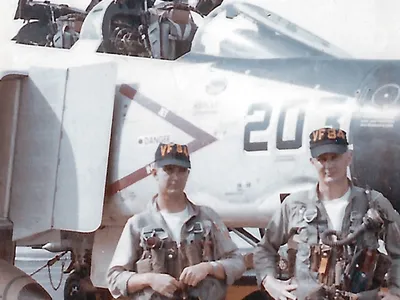 Two U.S. Navy men in uniform--a pilot and an aviator — stand in front of the cockpit of an F-4B Phantom II military jet aircraft.